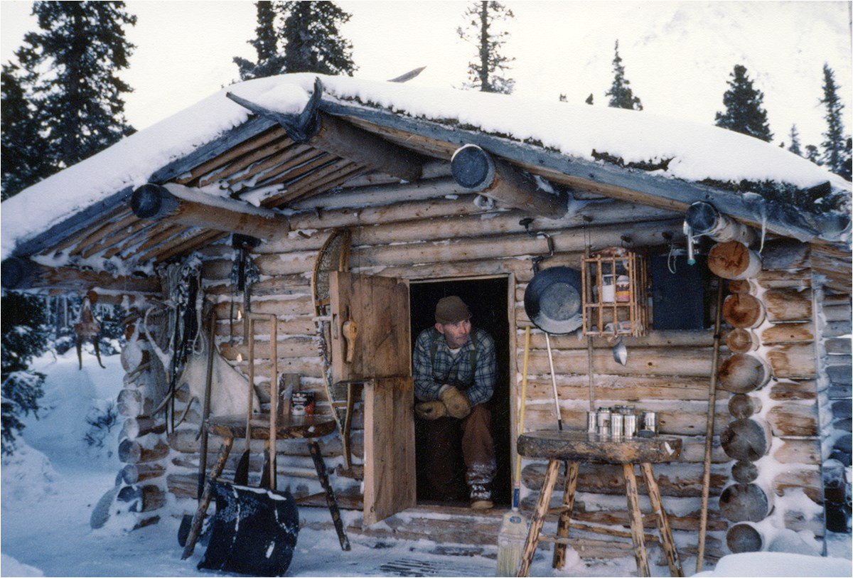 a man wearing a blue flannel shirt and brown pants sits in the open doorway of